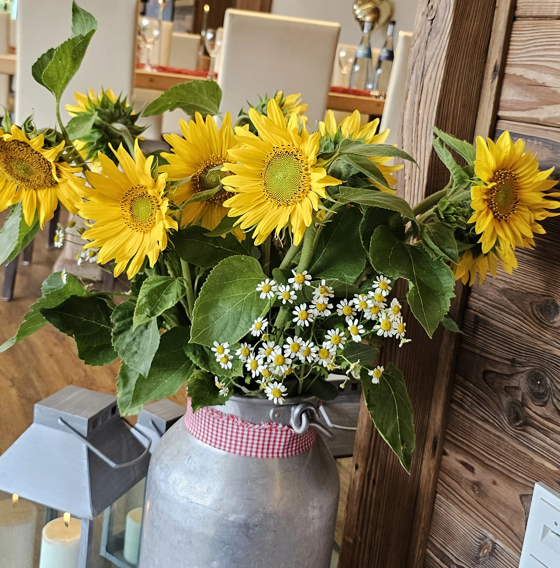 Ein silberner Milchkrug mit Sonnenblumen und kleinen weißen Blumen, auf einem Holztisch in einem rustikalen Raum.