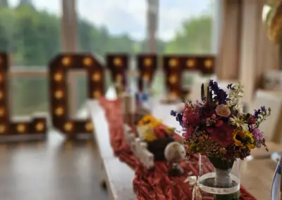 Tischdeko mit einem Blumenstrauß in einer Glasvase, im Hintergrund große, beleuchtete Buchstaben "LOVE". Rote Tischdecke und verschiedene Dekorationsartikel sichtbar.