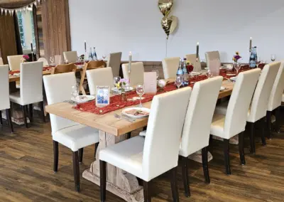 Ein festlich gedeckter Tisch in einem Restaurant mit weißen Stühlen und Holz-Tischplatten. Auf dem Tisch sind Gläser, Besteck und Blumenvasen sichtbar.