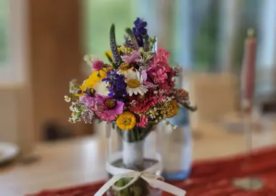 Bunte Blumenstrauß mit gelben, pinken und weißen Blumen in einer Glasvase, auf einem Tisch mit rotem Tischläufer.