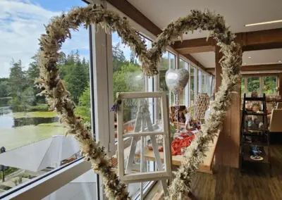 Ein herzförmiger Blumenkranz steht vor einem großen Fenster mit Blick auf einen See. Im Hintergrund sind Tische und Dekorationen für eine Hochzeit sichtbar.