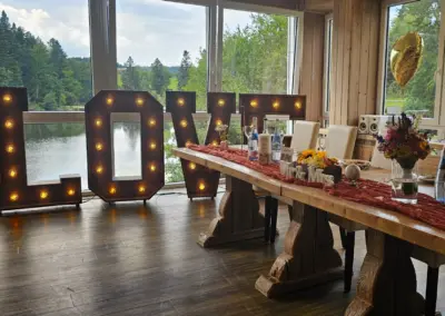 Großes, beleuchtetes 'LOVE'-Schild aus Holz steht neben einem festlich gedeckten Tisch mit Blumen, Flaschen und Gläsern, Blick auf einen See im Hintergrund.