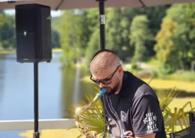 Ein Musiker spielt Saxophon auf einer Terrasse mit Blick auf einen See. Im Hintergrund sind Bäume und Wasser sichtbar.