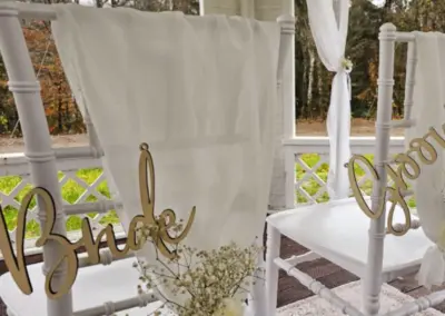 Zwei Stühle mit weißen Sitzflächen, einer beschriftet mit 'Bride' und der andere mit 'Groom'. Beide Stühle haben eine weiße Stoffbahn und eine kleine Blumendekoration.