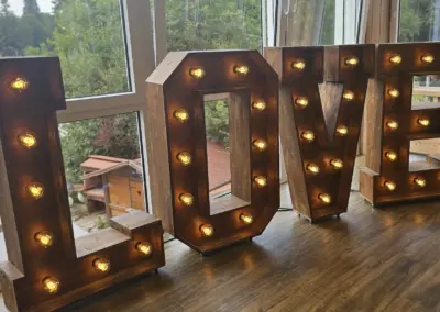 Holzbuchstaben 'L', 'O', 'V', 'E' mit Glühbirnen, auf einem Holzboden vor einem Fenster mit Blick auf Bäume.