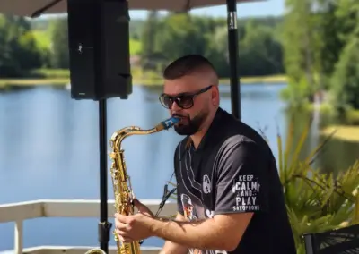 Ein Mann spielt Saxophon am Ufer eines Sees. Im Hintergrund sind Bäume und Wasser sichtbar. Er trägt ein schwarzes T-Shirt mit dem Text 'KEEP CALM AND PLAY SAXOPHONE'.