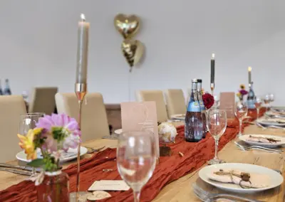 Festlich gedeckter Tisch für eine Hochzeit mit Kerzenhaltern, Gläsern, Wasserflasche und Blumen in kleinen Vasen. Ein goldener Herzballon schwebt im Hintergrund.