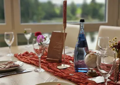 Tischdekoration für eine Hochzeit mit Gläsern, einer Flasche Wasser, einer Menükarte, Blumen und einem Globus auf einem Tisch mit Blick auf eine Landschaft.