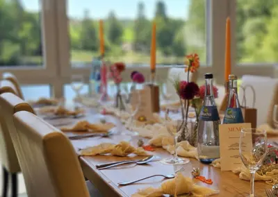 Ein festlich gedeckter Tisch mit weißen Stühlen, Gläsern, Flaschen, Kerzen und Blumenarrangements. Im Hintergrund ist eine grüne Landschaft sichtbar.