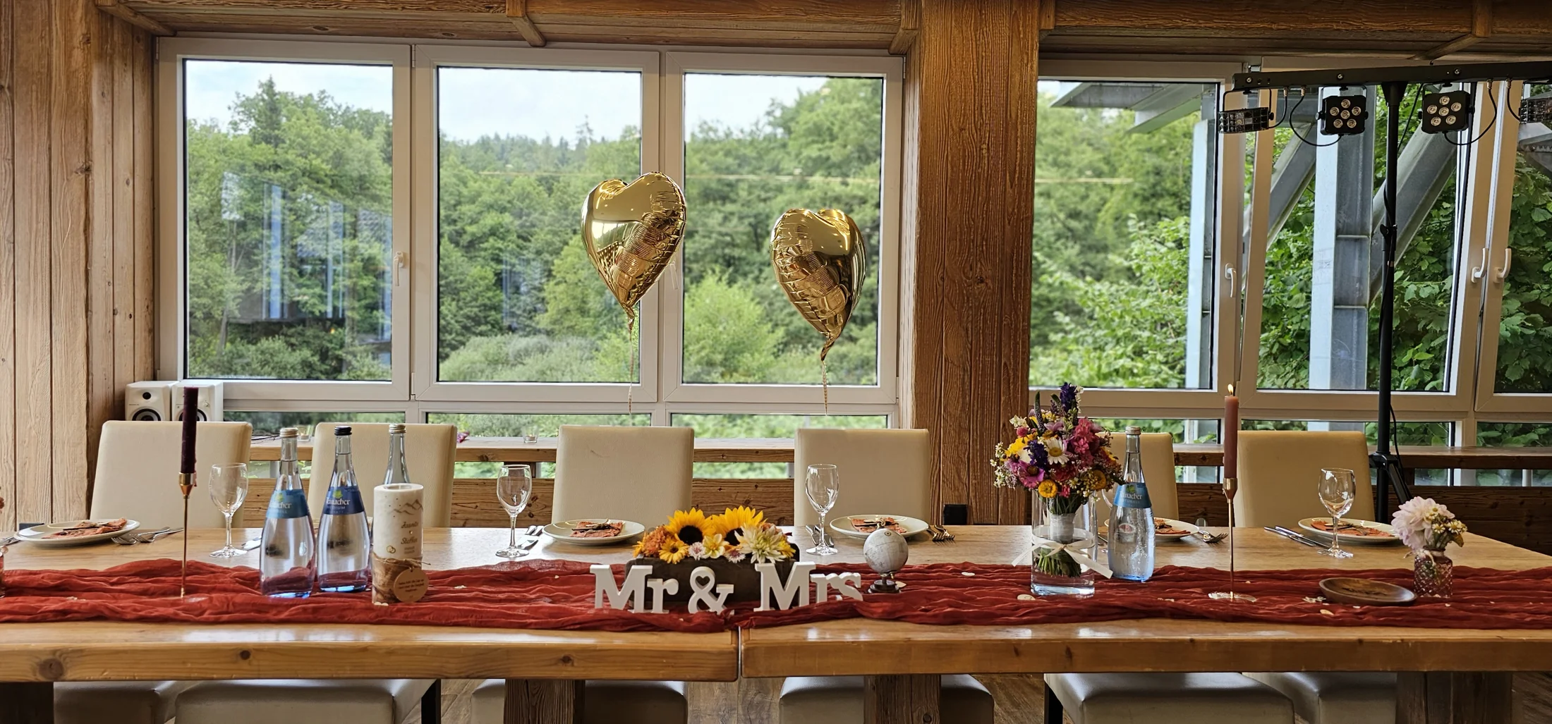 Ein festlich gedeckter Tisch für ein Hochzeitspaar mit goldenen Herzballons, Blumen, Gläsern und einer Tischdecke in Rot. Auf dem Tisch steht 'Mr & Mrs'.