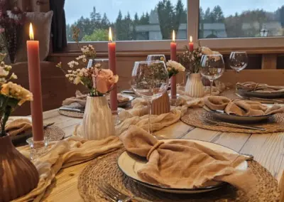 Festlich gedeckter Holztisch mit braunen Stoffservietten, Kerzen und kleinen Blumenvasen vor Fenster mit Waldsicht.
