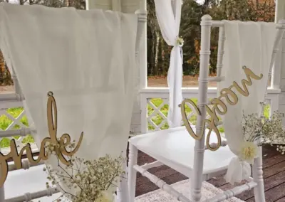 Zwei weiße Stühle mit weißen Stoffbezügen, beschriftet mit goldenen Holzschildern 'Bride' und 'Groom', dekoriert mit kleinen weißen Blumen.