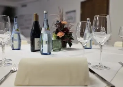Gedeckter Tisch mit mehreren Weingläsern, zwei Flaschen, Servietten und einem Blumenarrangement in einem Restaurant.