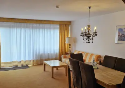 Wohnzimmer mit braunem Teppich, gelb-weiß gestreiftem Sofa, Holztisch, dunklen Stühlen, Kronleuchter und Treppe an der Wand.