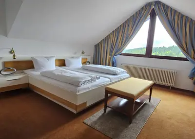 Hotelzimmer mit zwei Einzelbetten, Holzmöbeln, gelbem Teppich, Sessel und großem Fenster mit blauen Vorhängen.