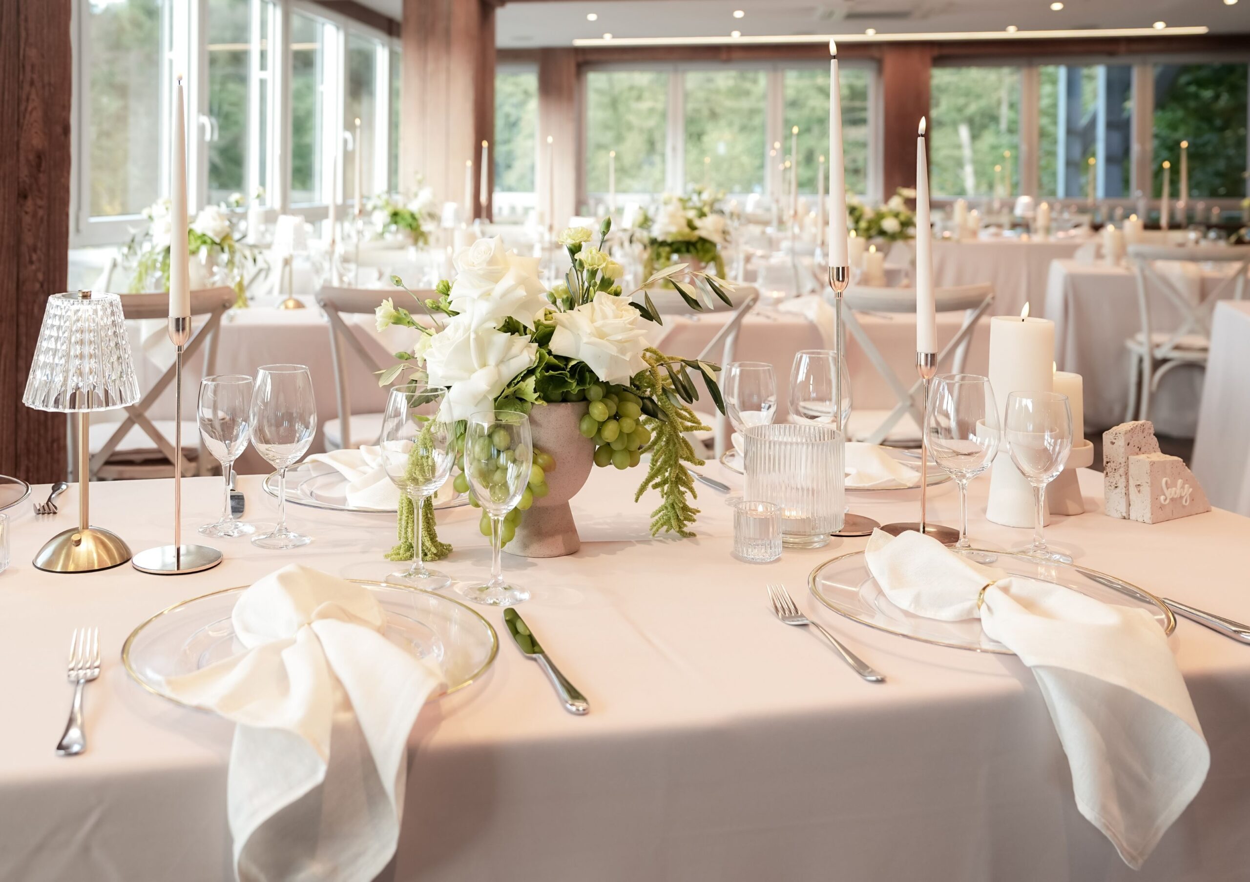 see_hotel_festliche_tafel_mit_seeblick_10 Festlich gedeckte Tische mit weißen Stühlen in einem Restaurant mit Blick auf einen Wald und einen See. Auf den Tischen stehen Gläser, Geschirr und Blumen.
