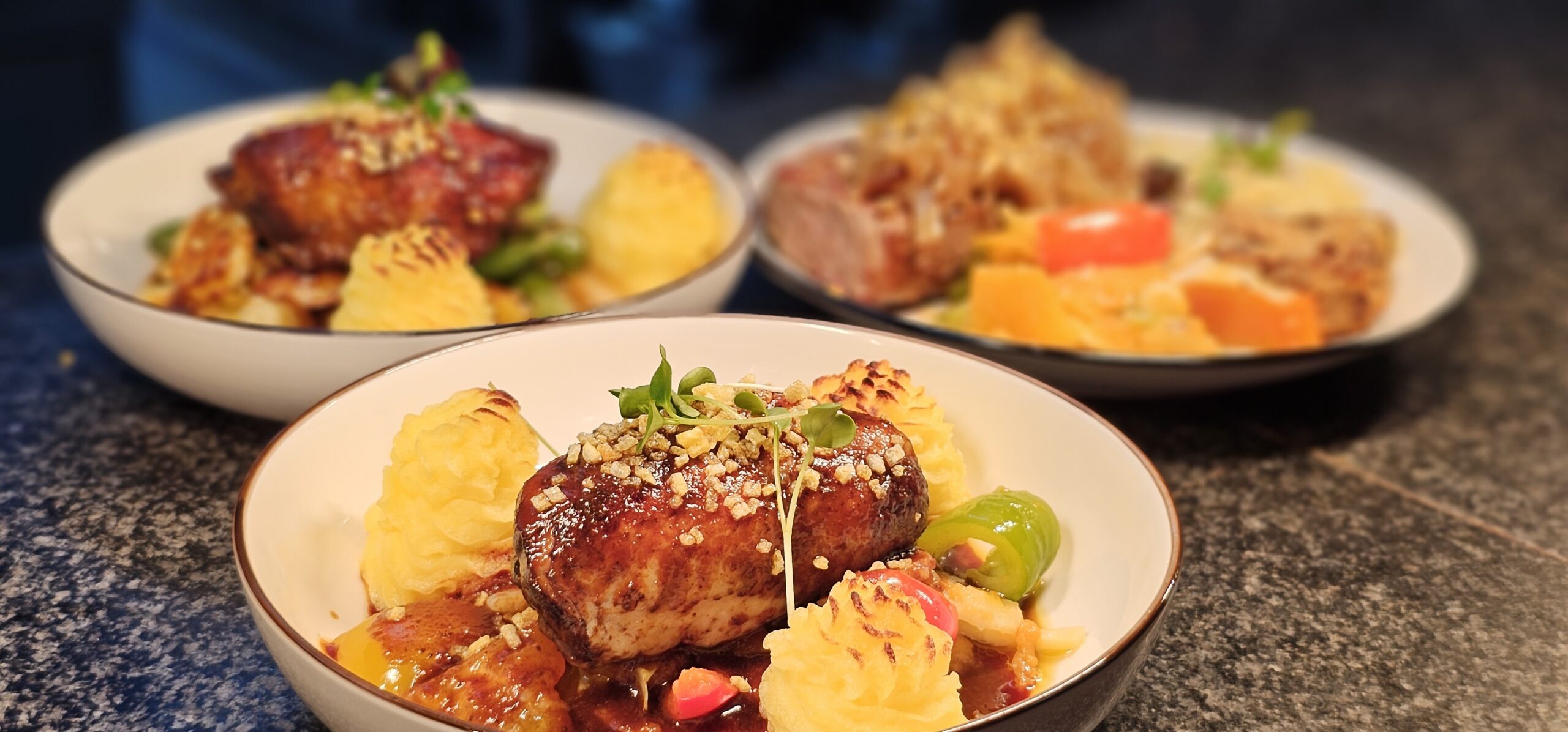 Reihe von fünf silbernen Buffet-Wärmeschalen auf einem Tisch, beschriftet mit 'Lamm', 'Hühnchen', 'Fisch', 'Gemüse' und 'Reis', vor einer Glasblockwand.
